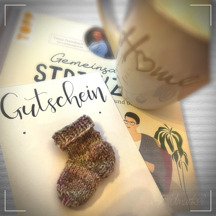 Buch, darauf eine Kaffeetasse, Gutschein mit einem kleinen, gestrickten Söckchen darauf. Präsentation einer Geschenkidee